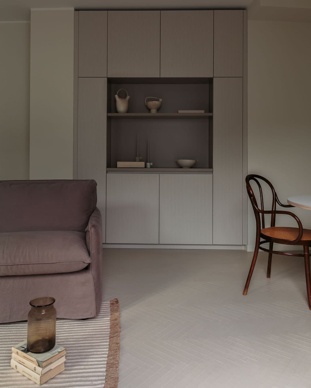 Cozy living room with a cozy sofa, wooden chair, and built-in shelves displaying pottery. A striped rug and glass vase adorn the floor.