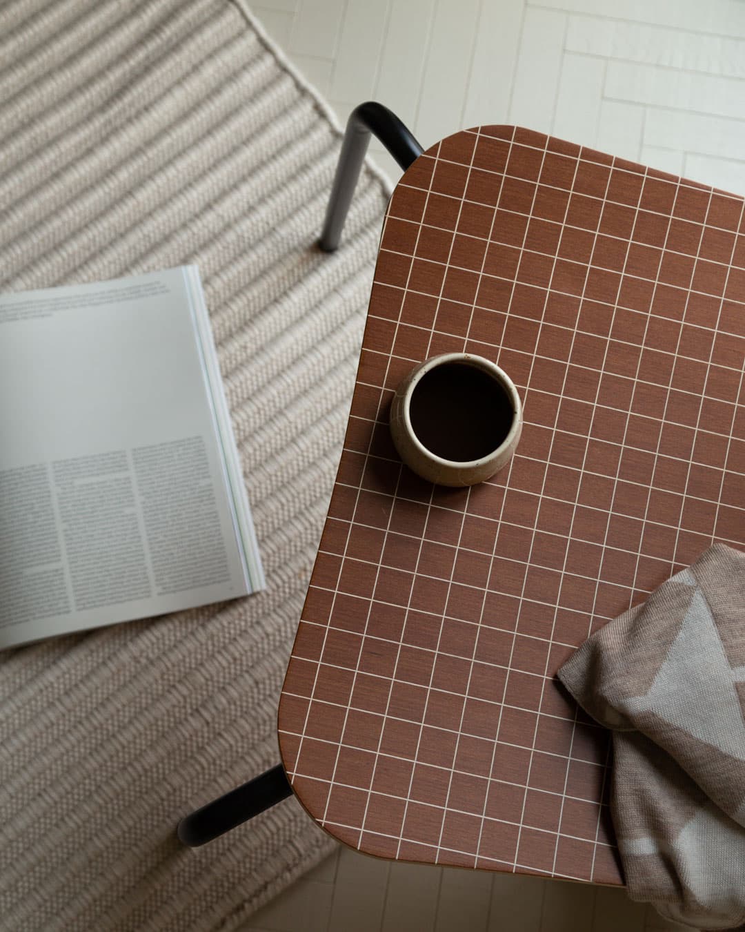 A brown grid-patterned table with a coffee cup, next to an open book on a textured rug.
