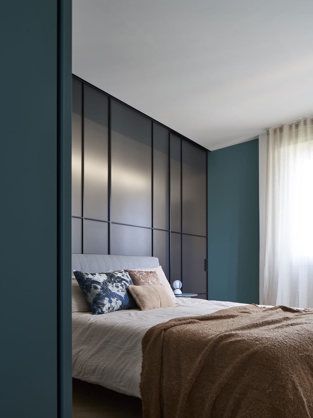 Modern bedroom with a sleek black accent wall, blue and beige pillows on a bed, and soft natural light filtering through sheer curtains.