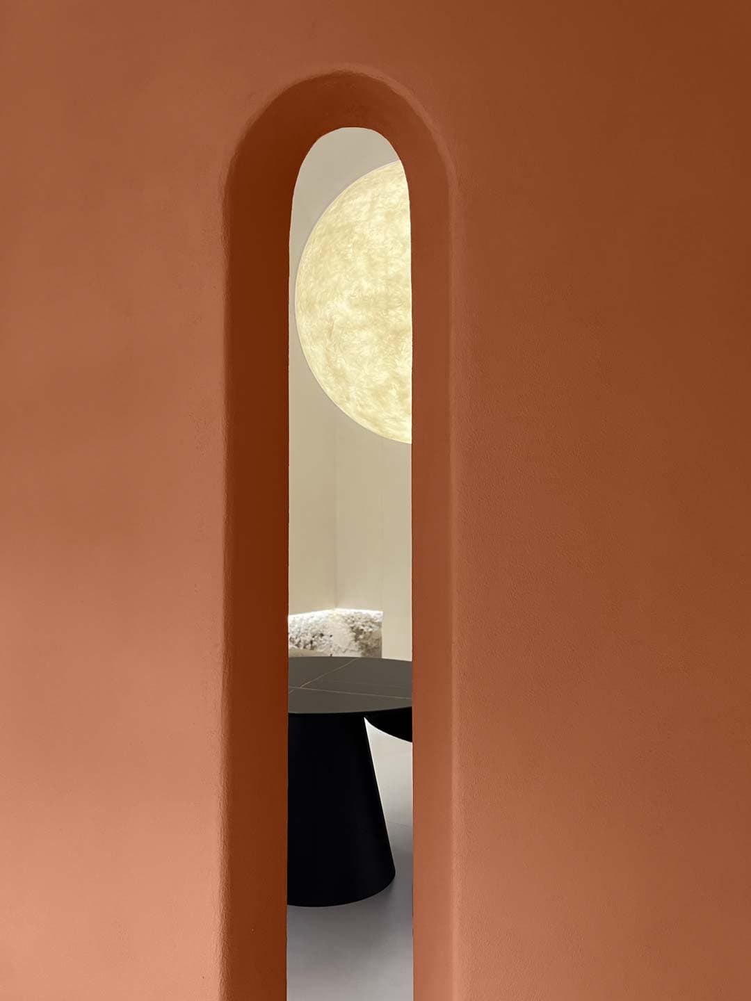A narrow, arched opening in a terracotta wall reveals a round table and a large, glowing circular light in the background.