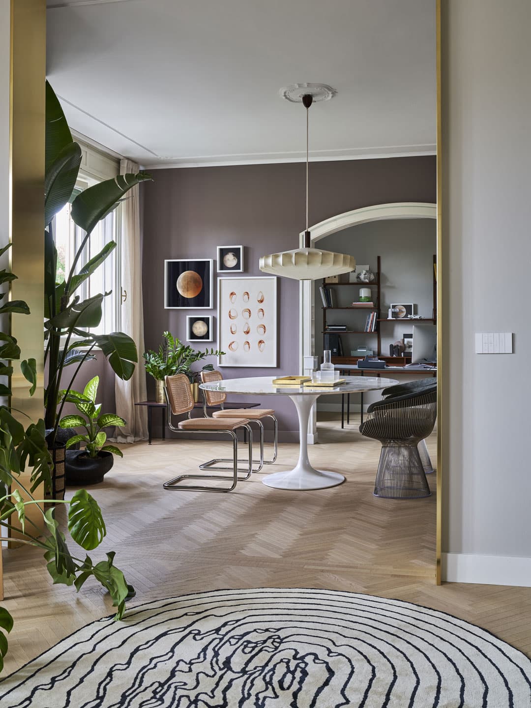 Modern dining area with a round table, chairs, large plants, and wall art. Neutral tones and natural light create a cozy atmosphere.