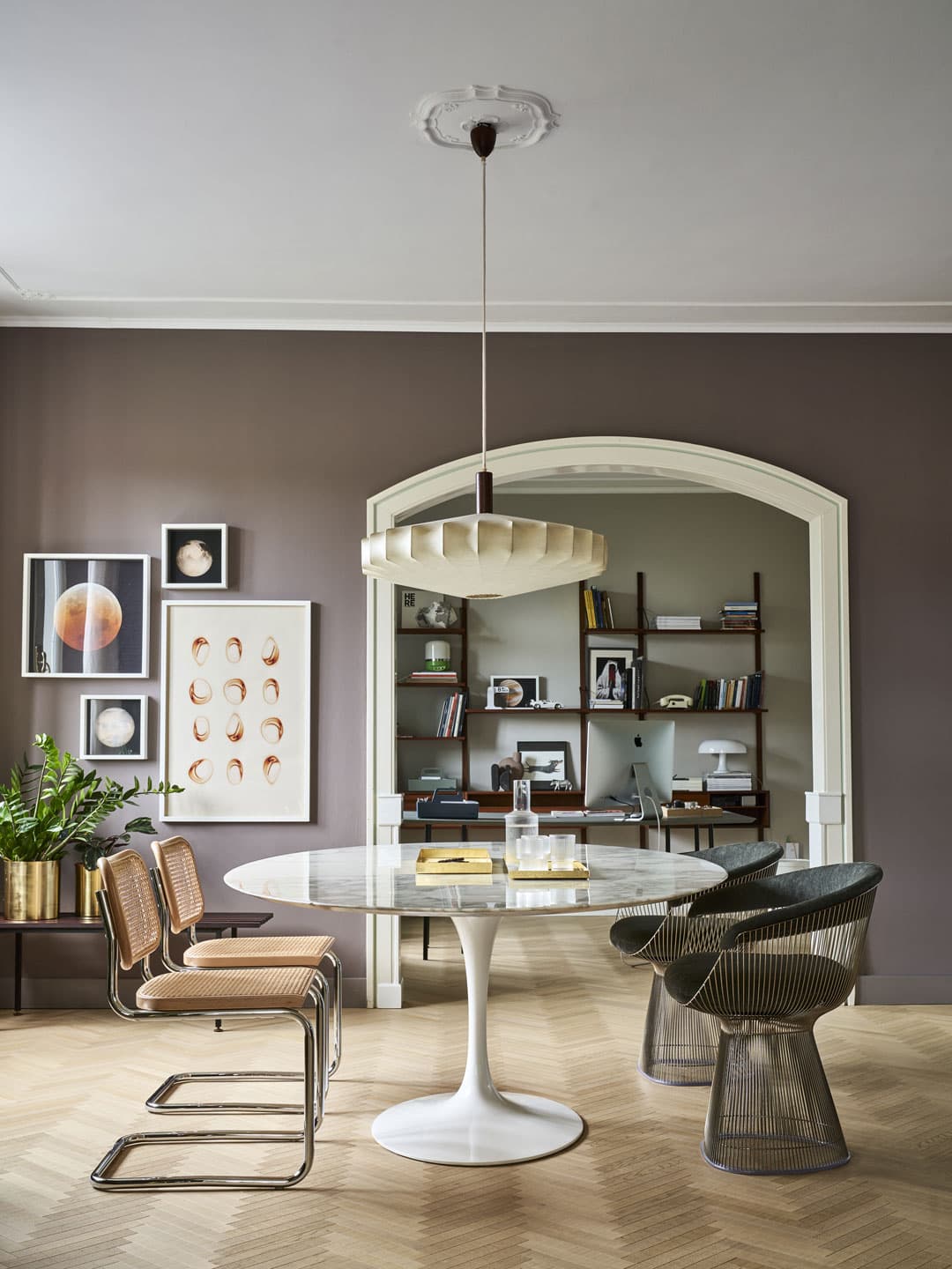 Modern dining room with a round marble table, mixed chairs, and a pendant light. Art on walls and bookshelves in the background.