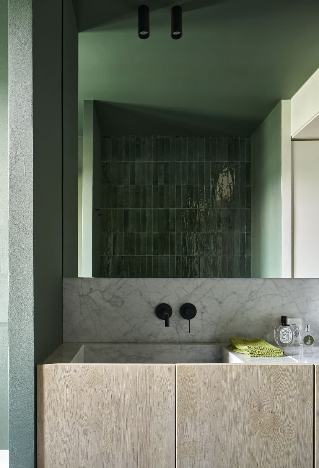 Modern bathroom with green tiles, a large mirror, wood vanity, marble countertop, black fixtures, and a folded green towel beside toiletries.