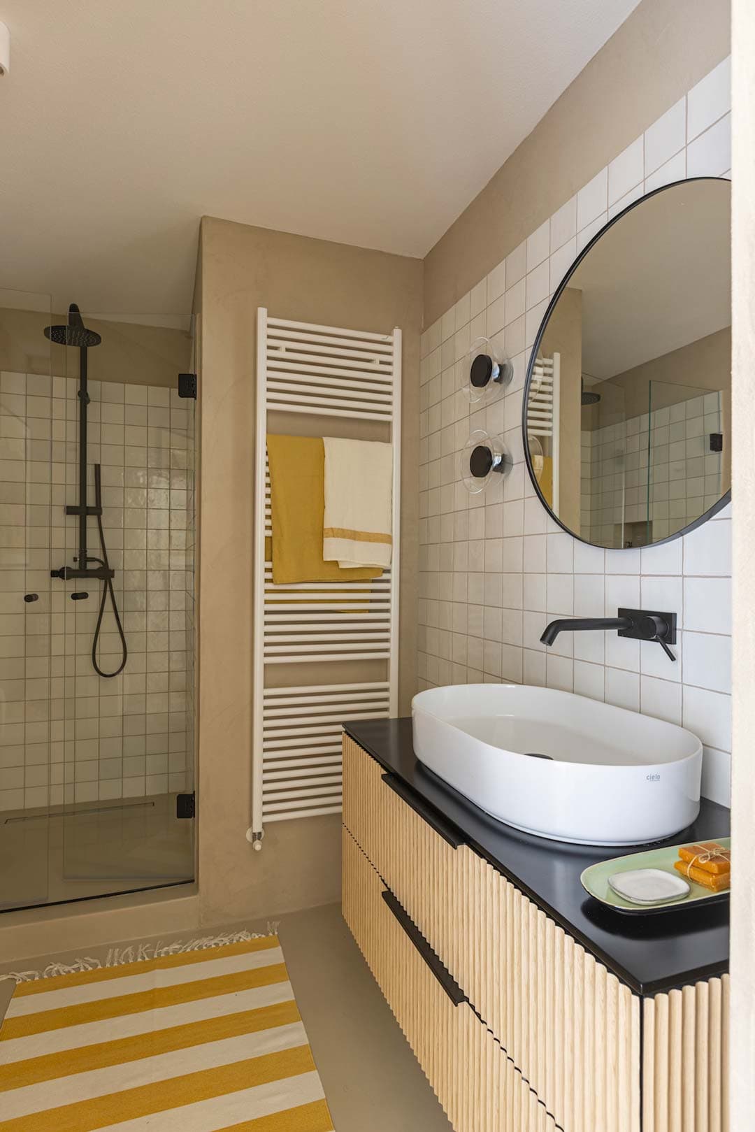 Modern bathroom with a round mirror, white sink on a wooden vanity, striped rug, glass shower, and towel rack with yellow and white towels.