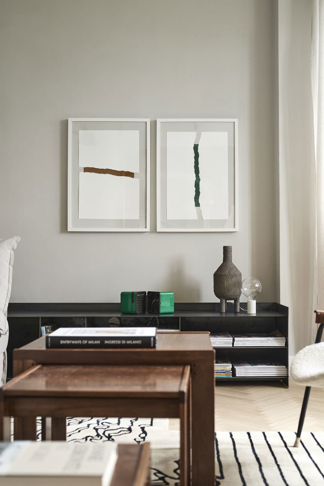 Minimalist living room with two framed abstract artworks on the wall, a low shelf with books and decor, and a wooden coffee table on a patterned rug.