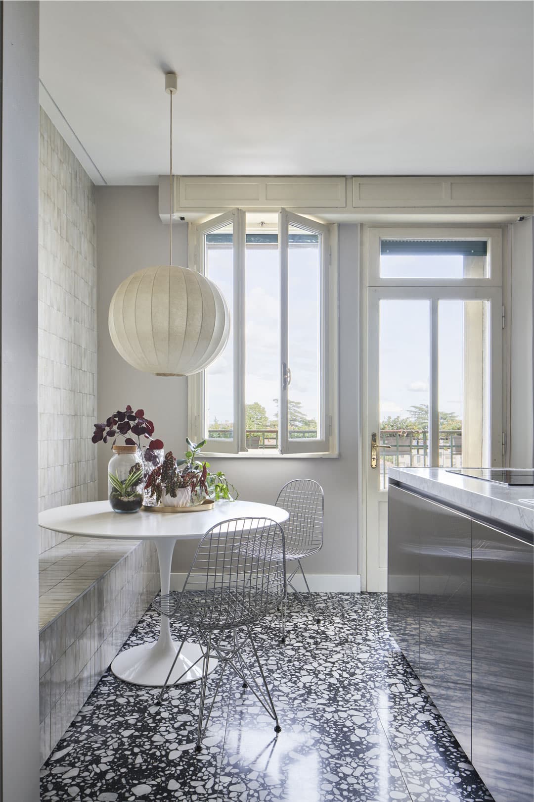 Modern kitchen with terrazzo floor, round white table, wire chairs, and a large pendant light. Large windows offer a view outside.