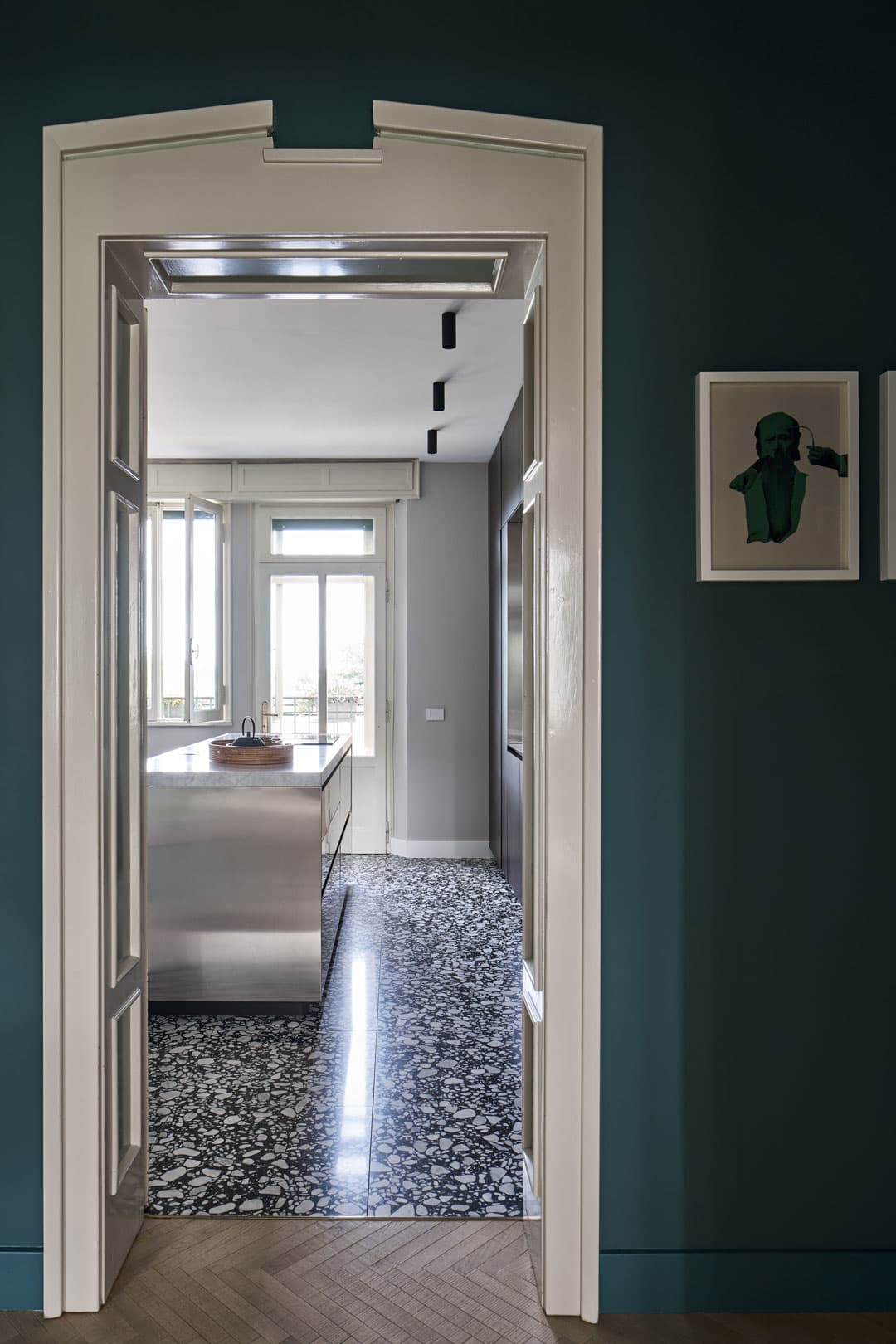 Doorway view into a modern kitchen with a terrazzo floor, stainless steel island, large windows, and teal walls with a small framed artwork.