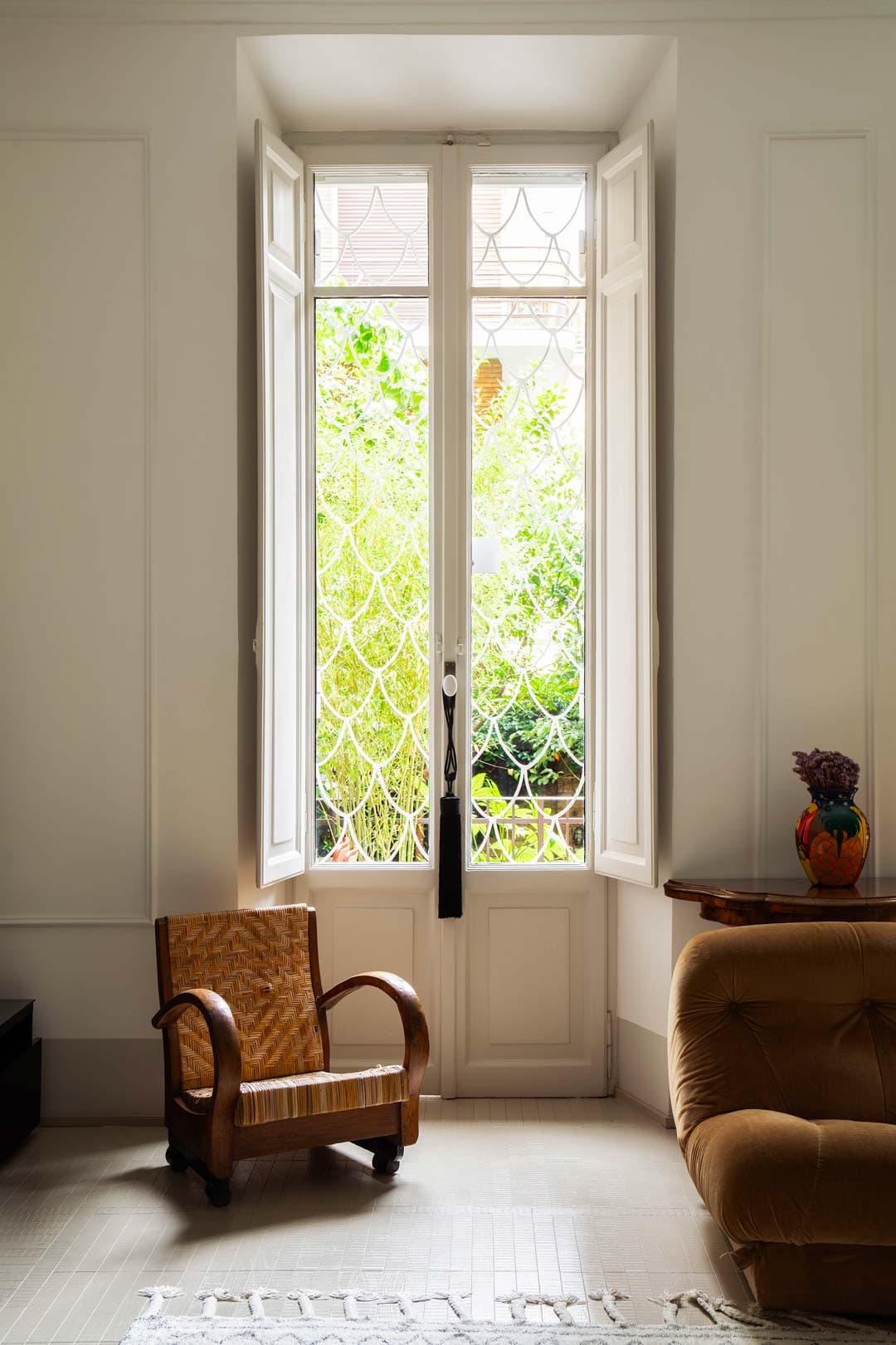 A cozy room with a wicker chair and brown sofa, featuring a large window with intricate metalwork, overlooking a lush garden.