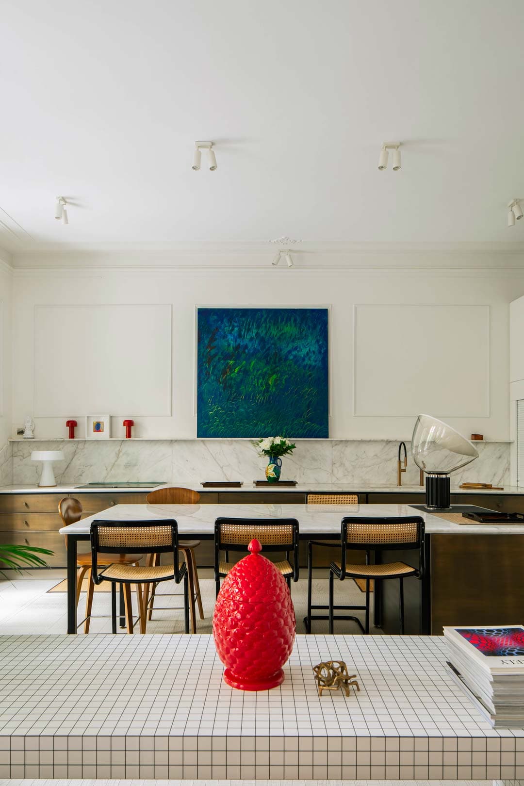 Modern kitchen with a large marble island, wooden stools, red decorative vase, and a vibrant blue-green painting on the wall.