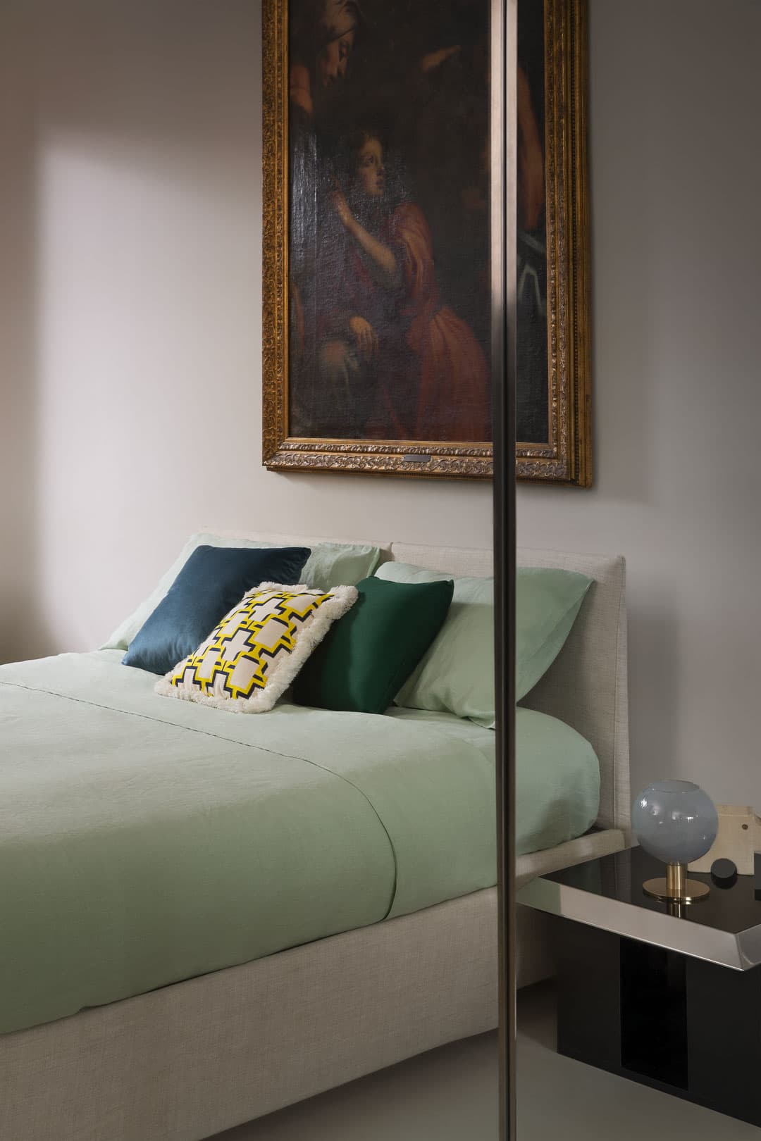 Bedroom with a light green bed, decorative pillows, a side table with a globe lamp, and a large framed painting above the headboard.