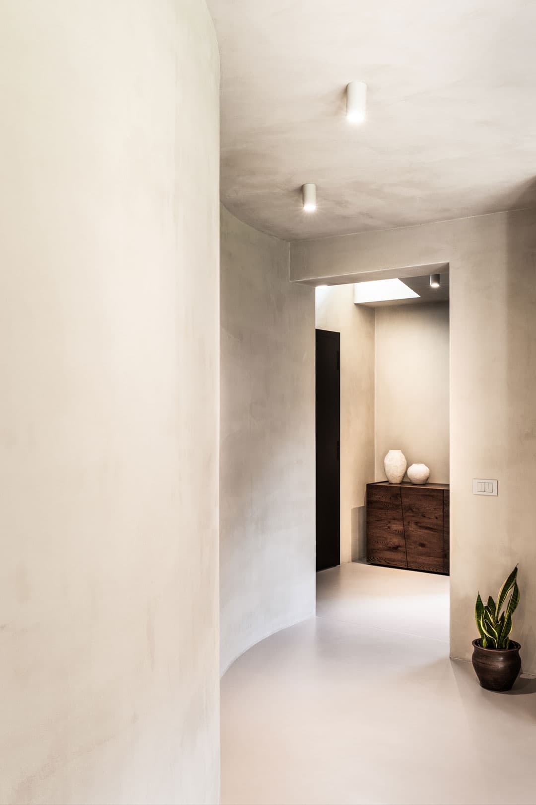 Minimalist hallway with curved beige walls, a wooden cabinet with decorative vases, and a potted plant in the corner.