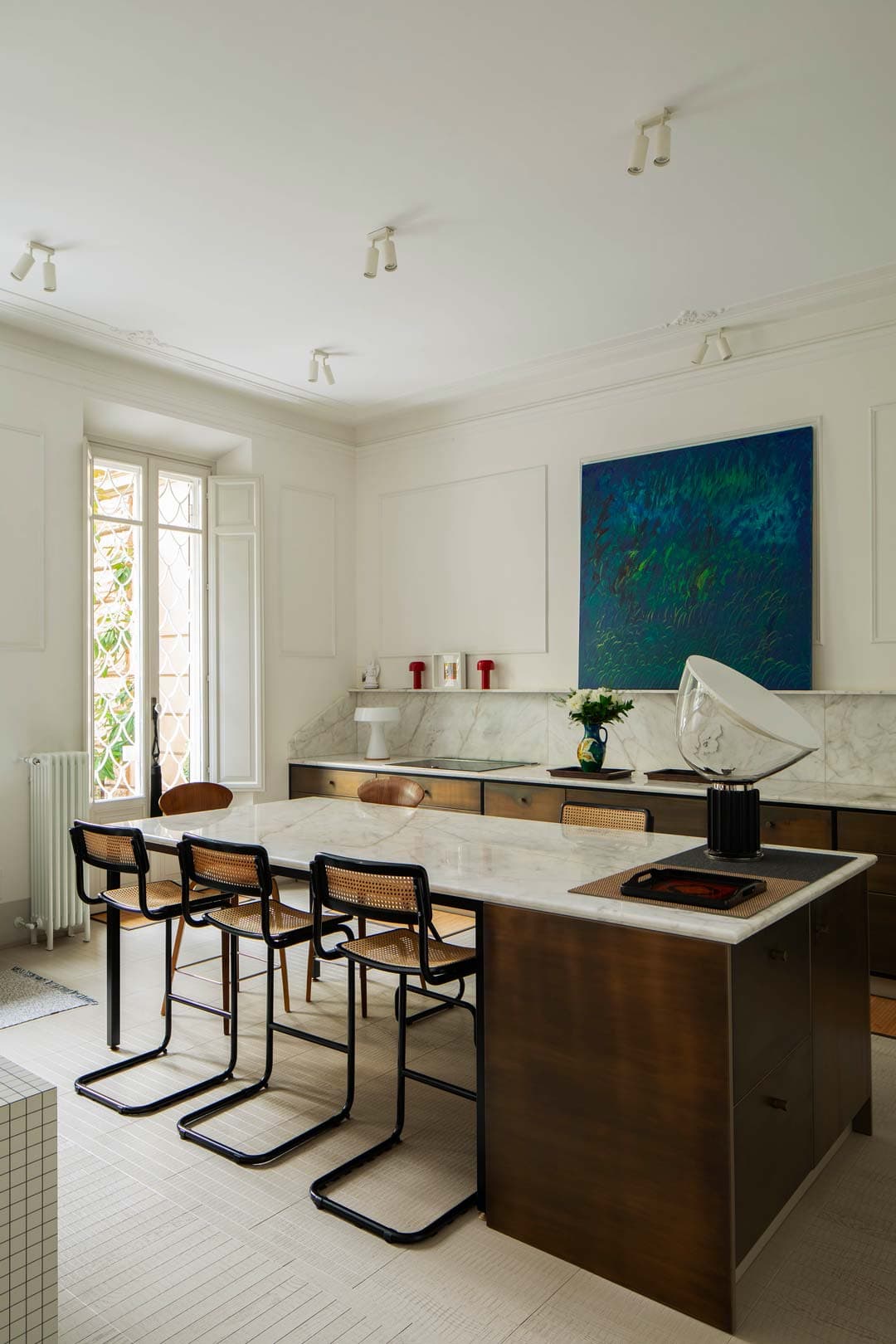 Modern kitchen with a large marble island, wooden cabinets, and cane-backed chairs. A blue abstract painting and sculpture decorate the space.