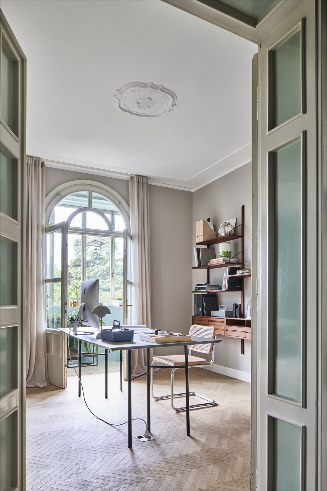 A home office with a desk, computer, and chair by a large arched window. Shelves hold books and decor. Light wood herringbone floor.