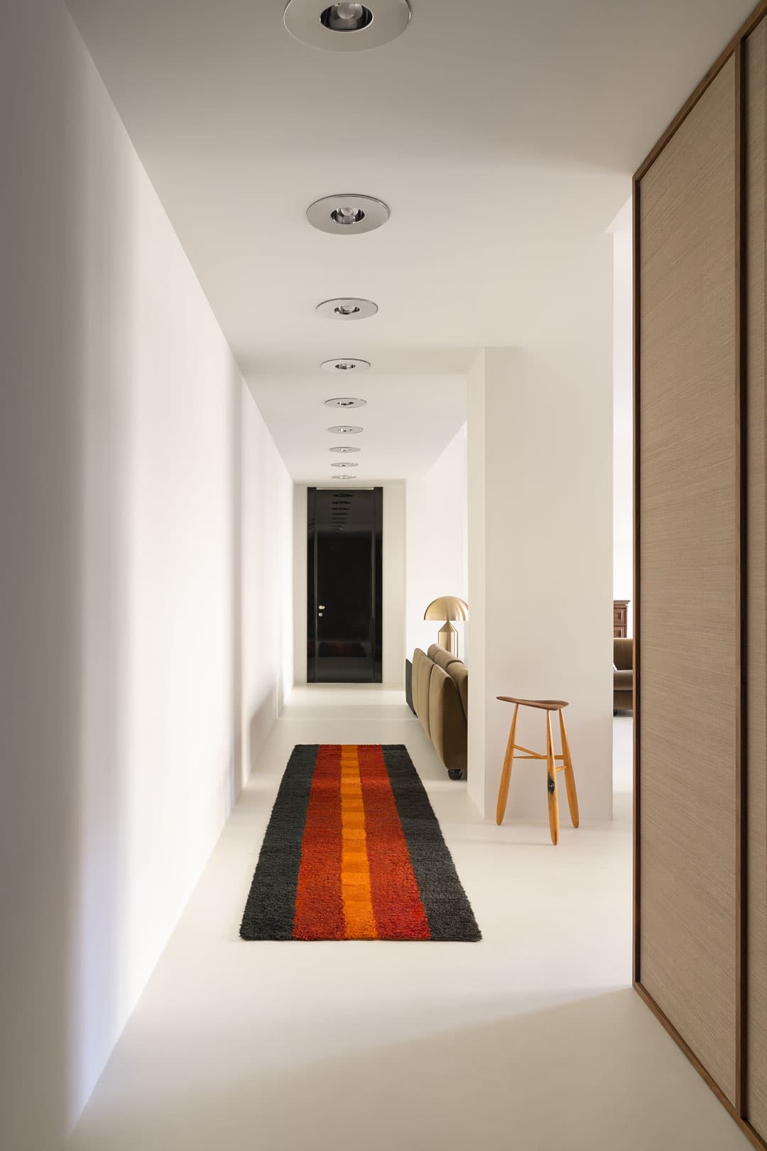 Minimalist hallway with white walls, recessed lighting, a striped rug, and a wooden stool. A sofa and lamp are visible at the end.