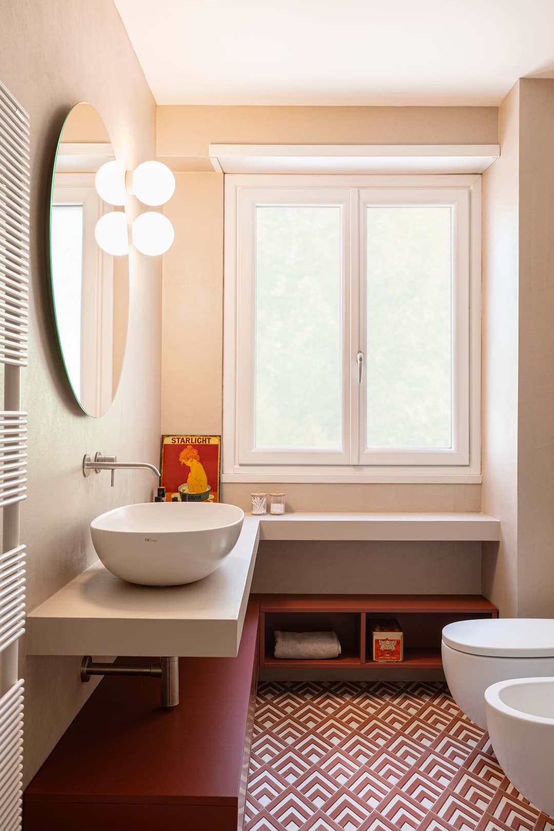 Modern bathroom with round mirror, white sink, and geometric patterned floor. A window lets in natural light, and shelves hold essentials.
