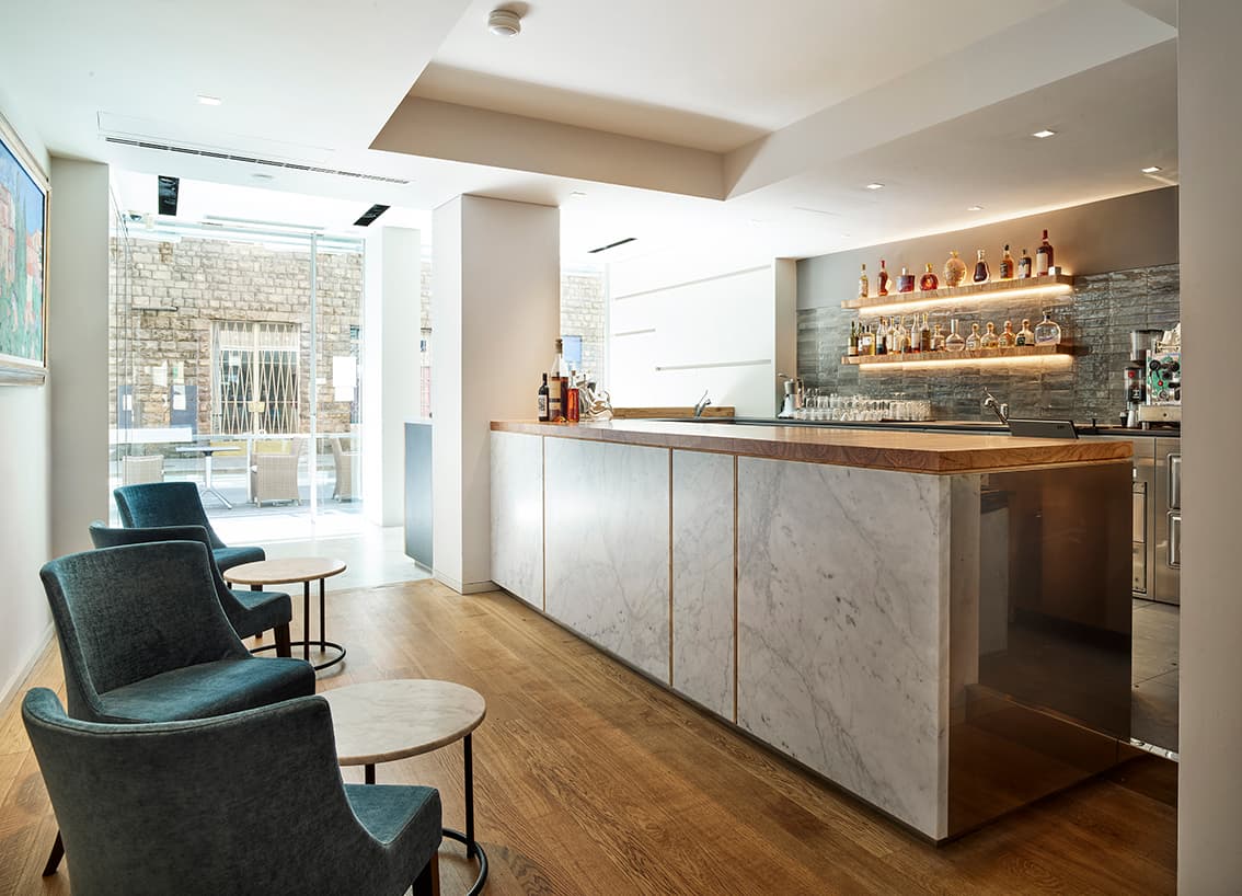 Modern bar interior with a marble counter, wooden floor, blue chairs, and a well-lit shelf displaying bottles. Large window in the background.