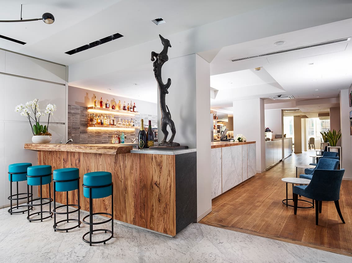 Modern hotel lobby with a wooden bar, teal stools, a metal sculpture, and a seating area with blue chairs on a wooden floor.