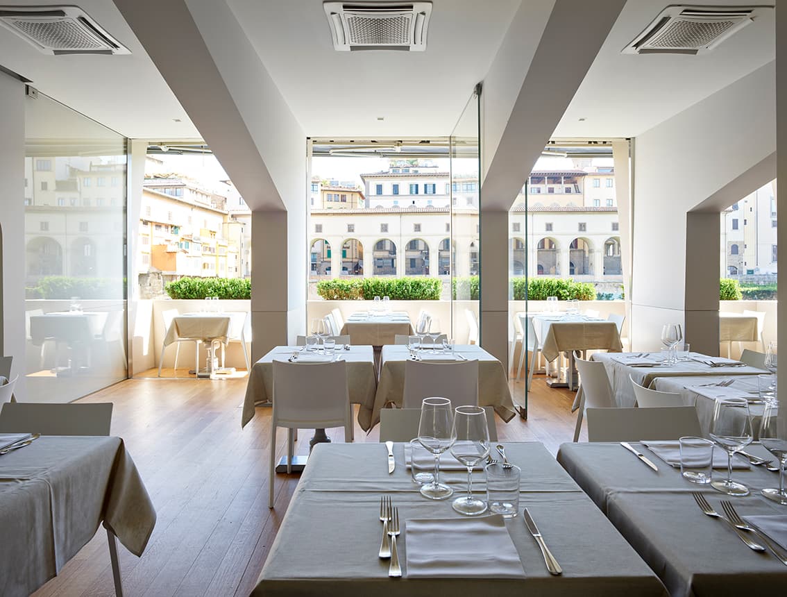 Modern restaurant interior with neatly set tables, white tablecloths, and large windows overlooking a cityscape with historic buildings.