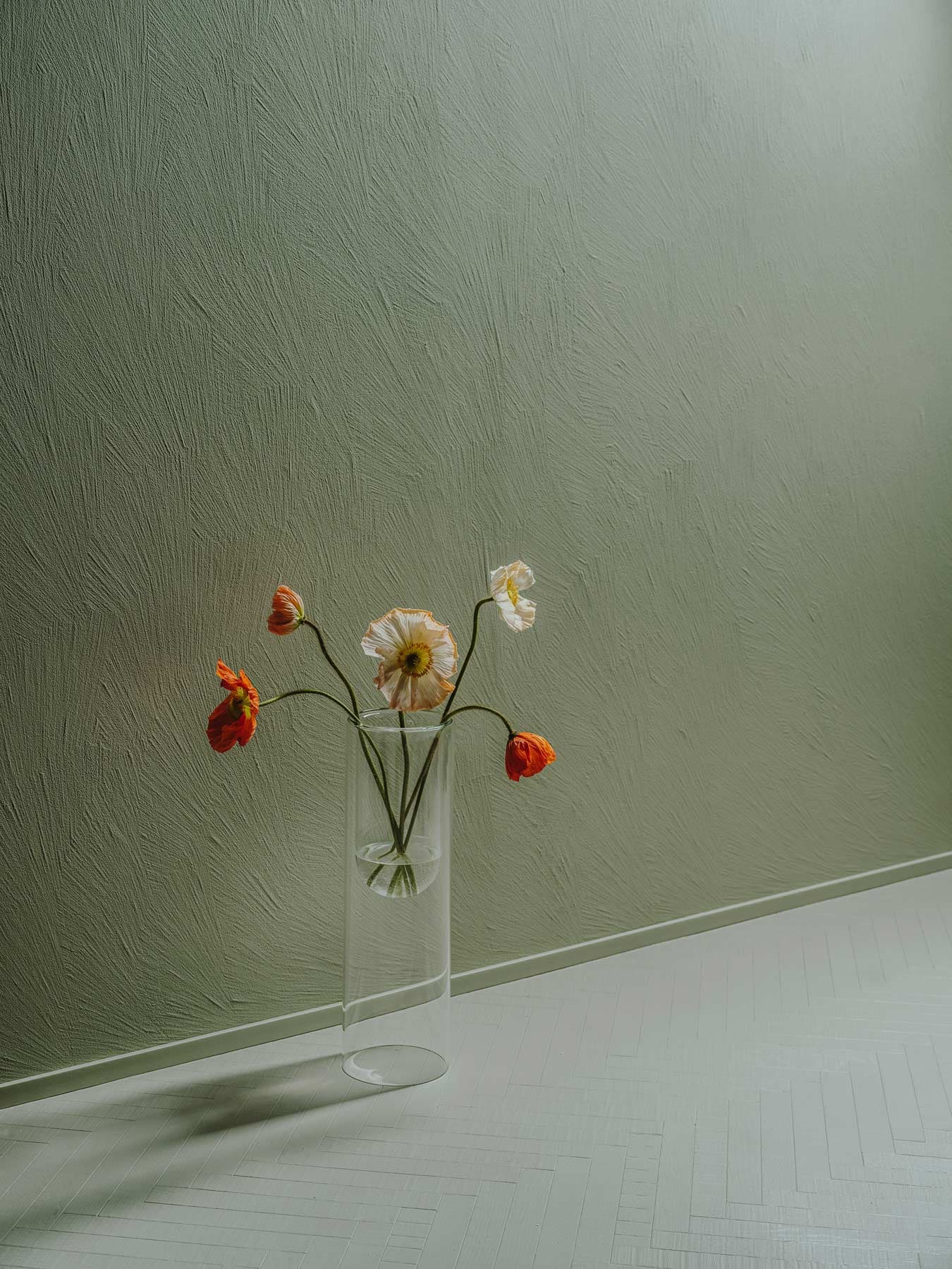 A clear vase with red and white flowers against a textured green wall on a light floor.