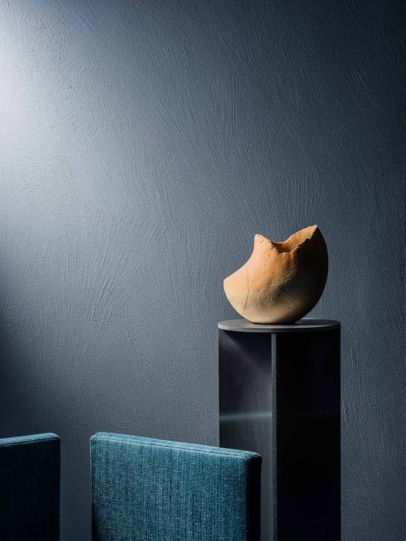 A cracked, round clay vase sits on a tall, dark pedestal against a textured gray wall, with part of a blue chair visible in the foreground.