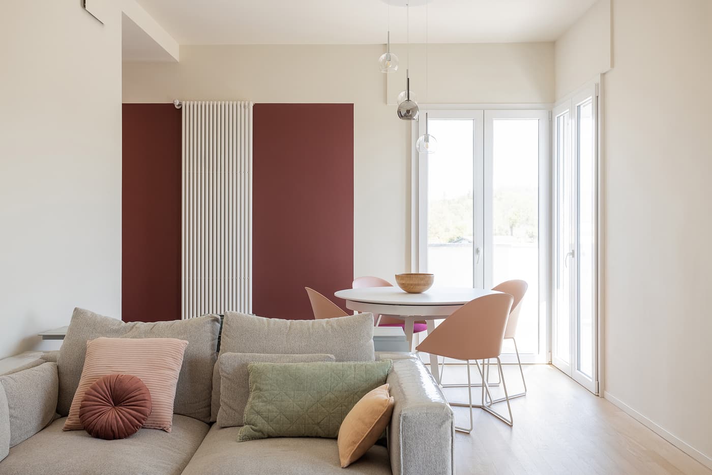 Modern living room with a beige sofa, colorful cushions, round dining table, pink chairs, and large windows with natural light.
