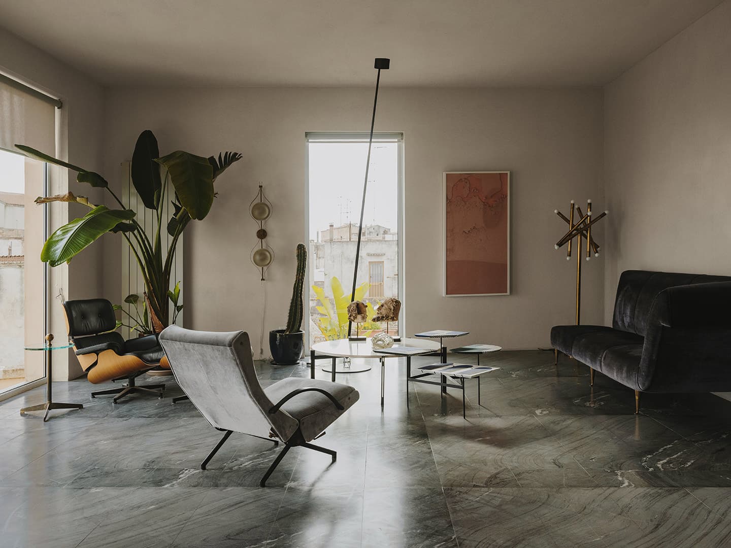 Modern living room with stylish chairs, a black sofa, large potted plants, abstract art, and a floor lamp on a marble floor.