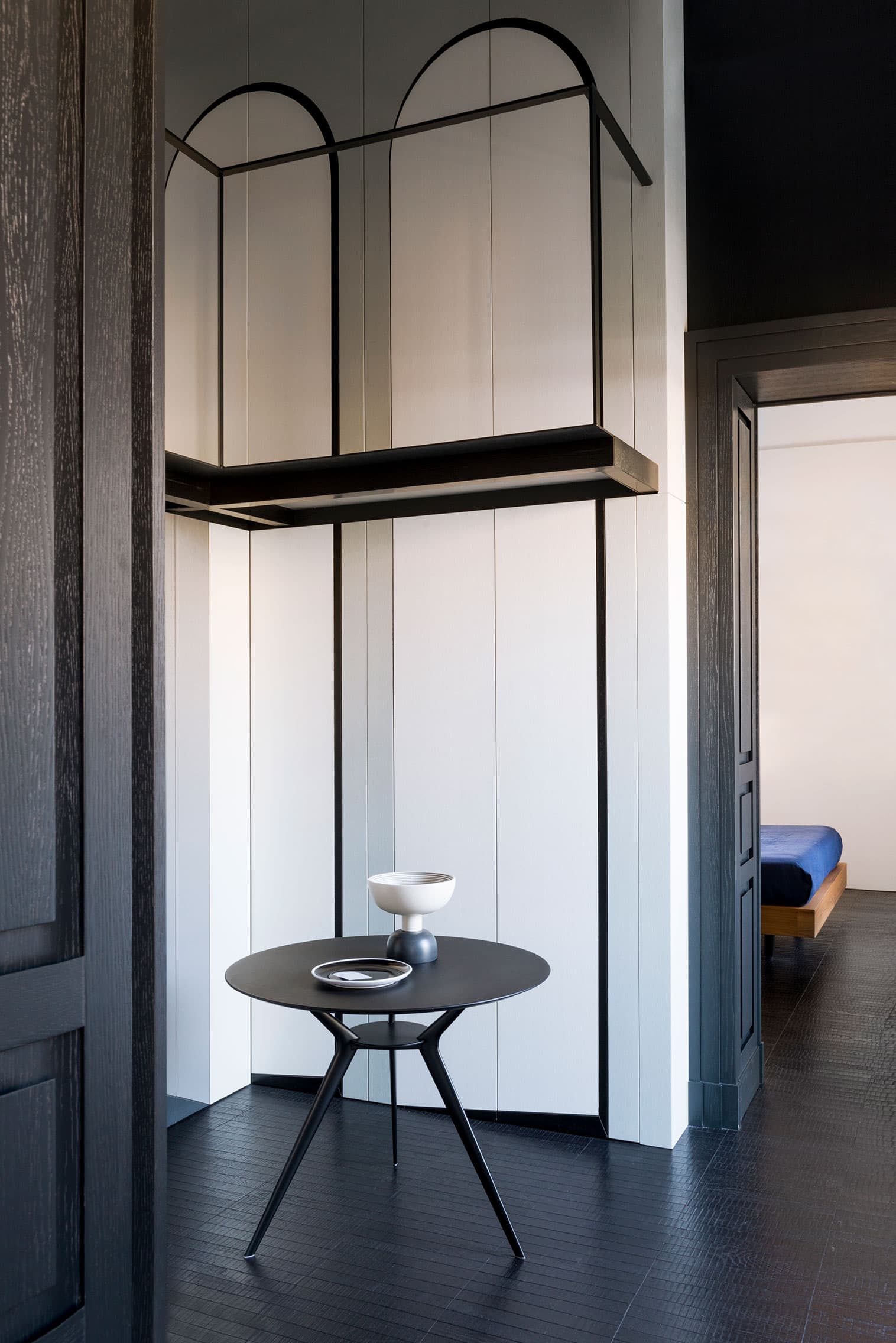 Minimalist room with a black round table, and a black shelving unit against white panel walls. A bedroom is visible in the background.