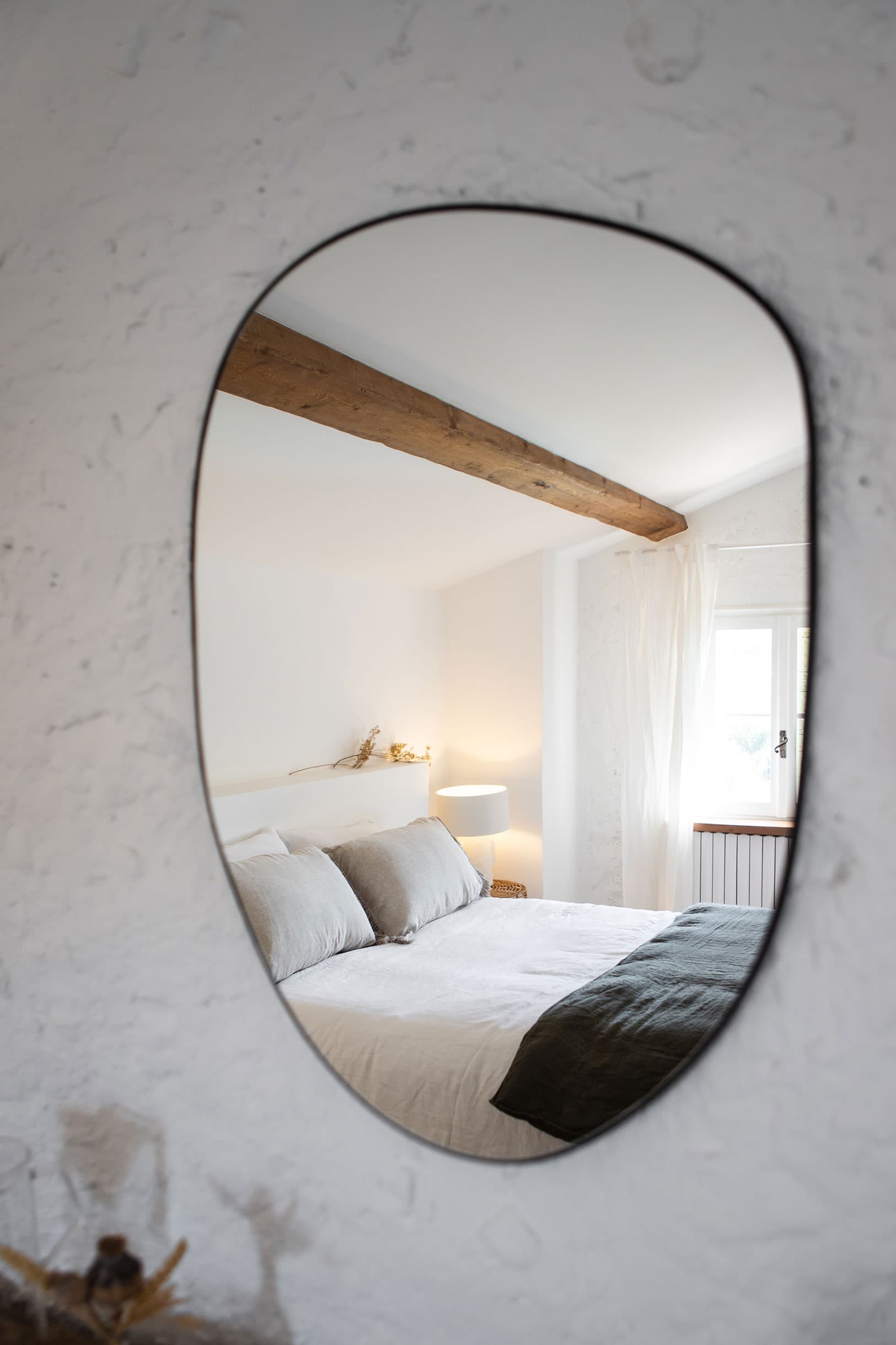 A cozy bedroom reflected in an oval mirror, featuring a wooden beam, white bedding, gray pillows, a bedside lamp, and a window with sheer curtains.