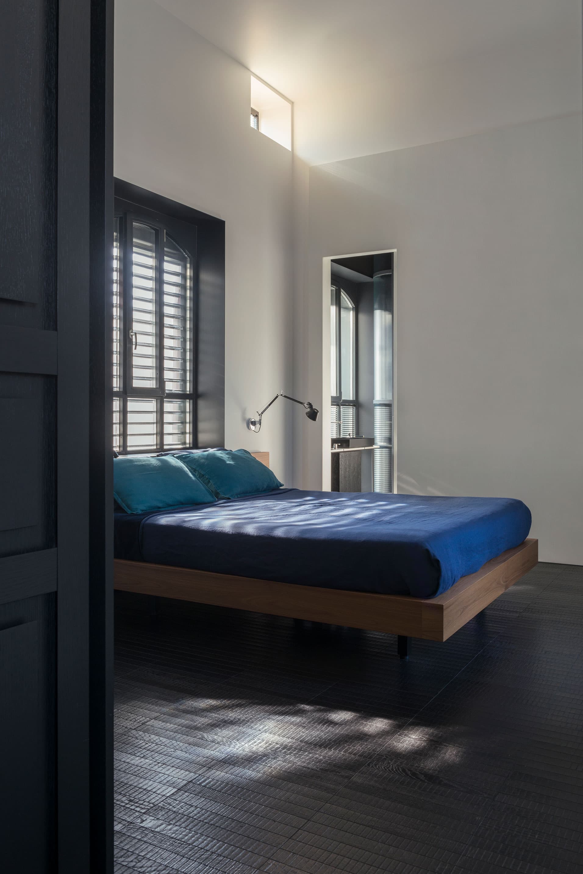 Minimalist bedroom, a bed with blue bedding, dark floors, and a wall-mounted lamp. Sunlight streams through shuttered windows.