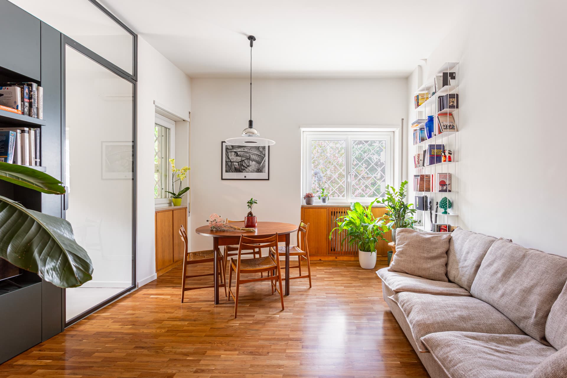 Living room with wooden floor, cozy beige sofa, round dining table, pendant light, and large plants near a window with bookshelves.