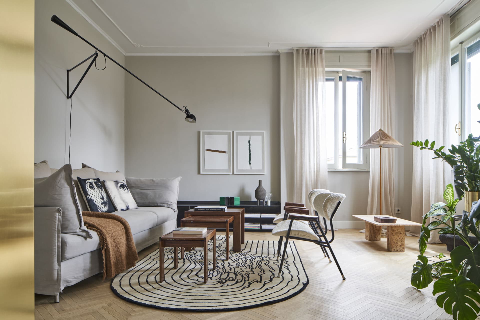 Modern living room with a beige sofa, patterned rug, wooden tables, abstract art, and large windows with sheer curtains.
