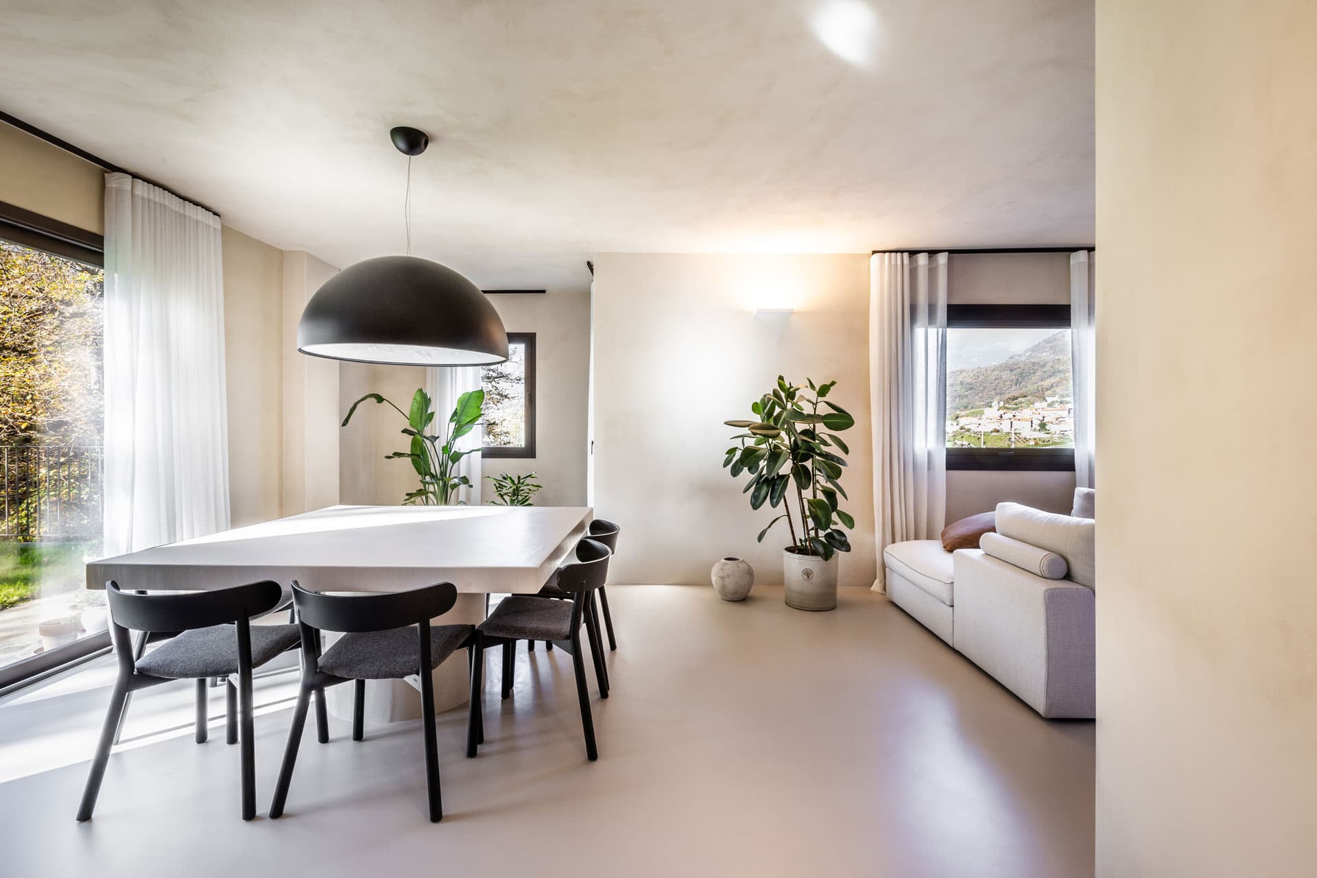 Modern dining room with a large white table, black chairs, pendant light, and potted plants. Large windows provide natural light and a scenic view.