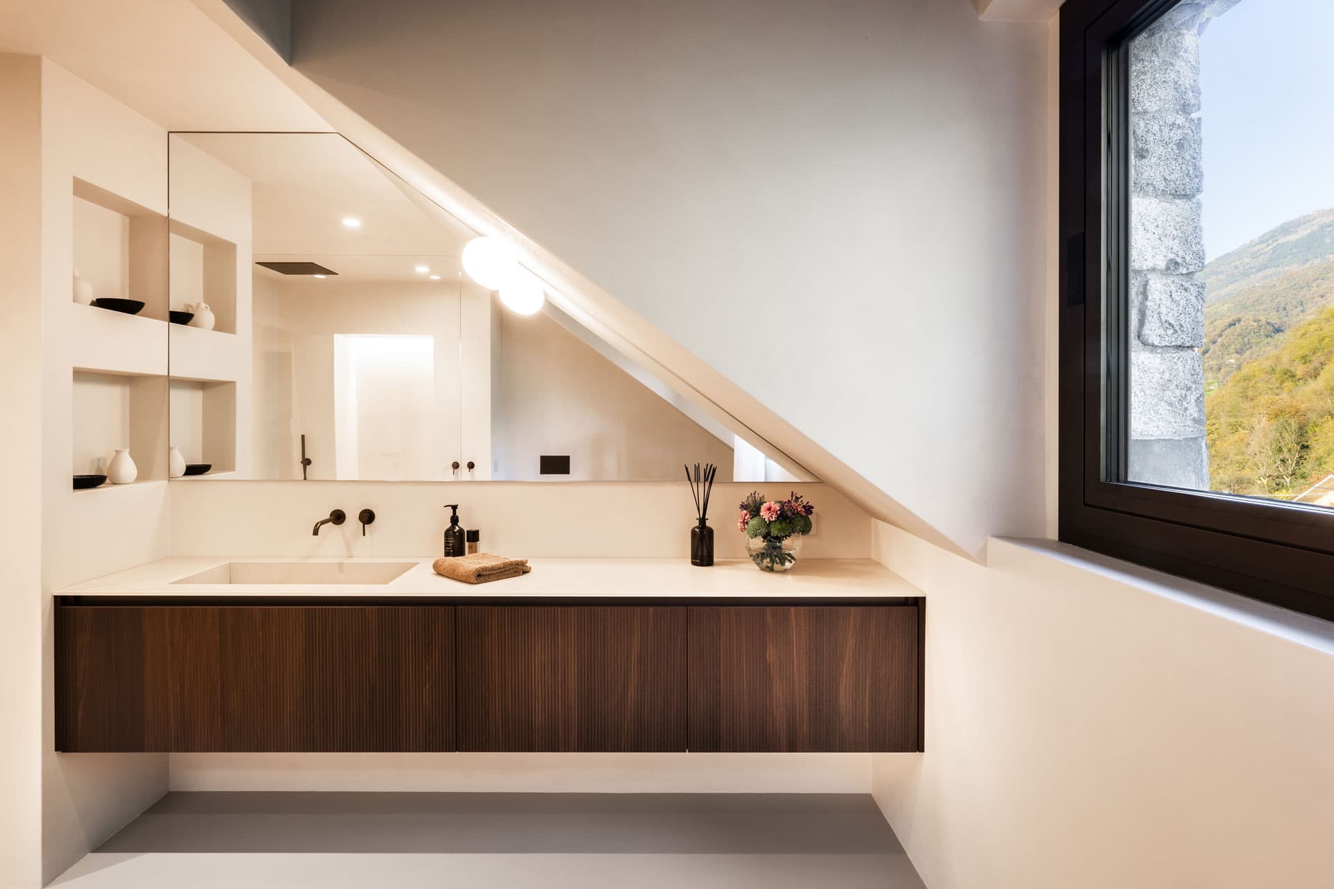 Modern bathroom with sloped ceiling, large mirror, wooden vanity, and window with a scenic view. Decor includes flowers and toiletries.
