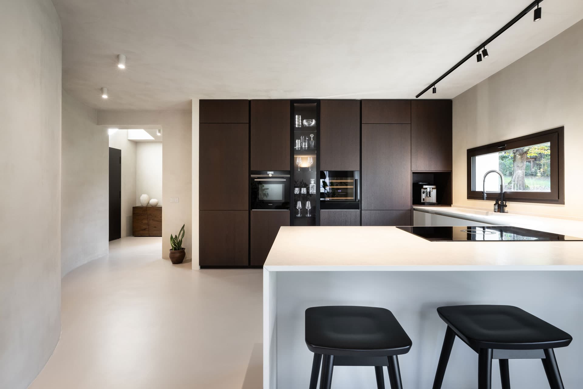 Modern kitchen with dark wood cabinets, built-in appliances, a sleek island with a cooktop, and two black stools. Natural light from a large window.