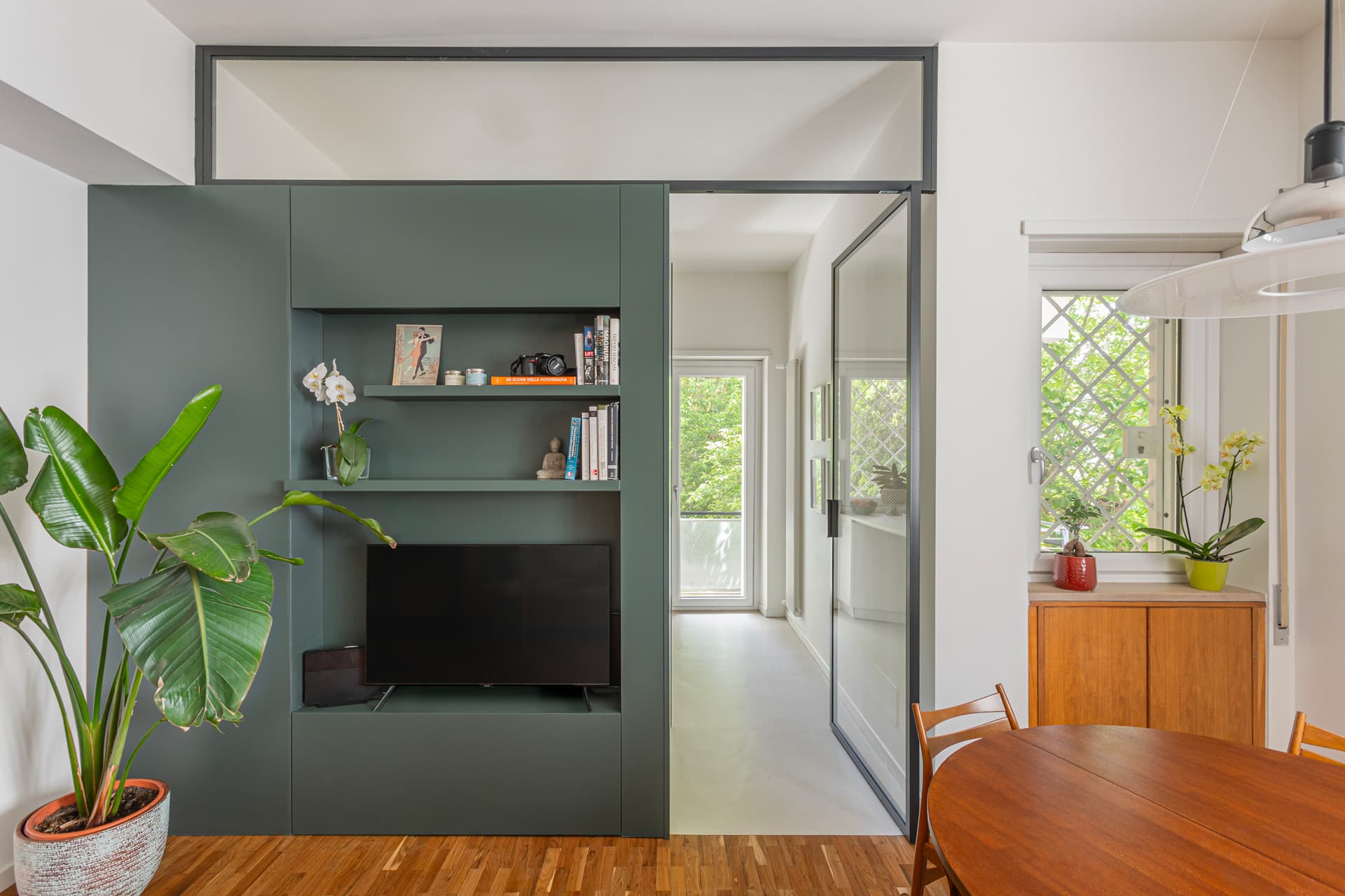 Modern living space with green built-in unit, TV, books, and plants. Wooden dining table and chairs, large windows, and bright natural light.