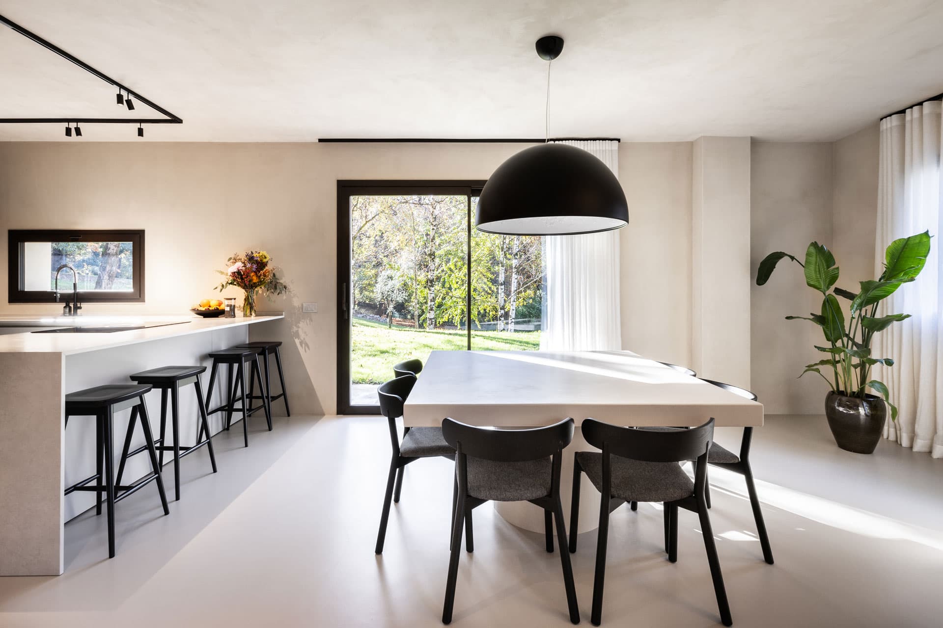 Modern dining area with a large white table, black chairs, a black pendant light, and a view of a garden through a sliding glass door.