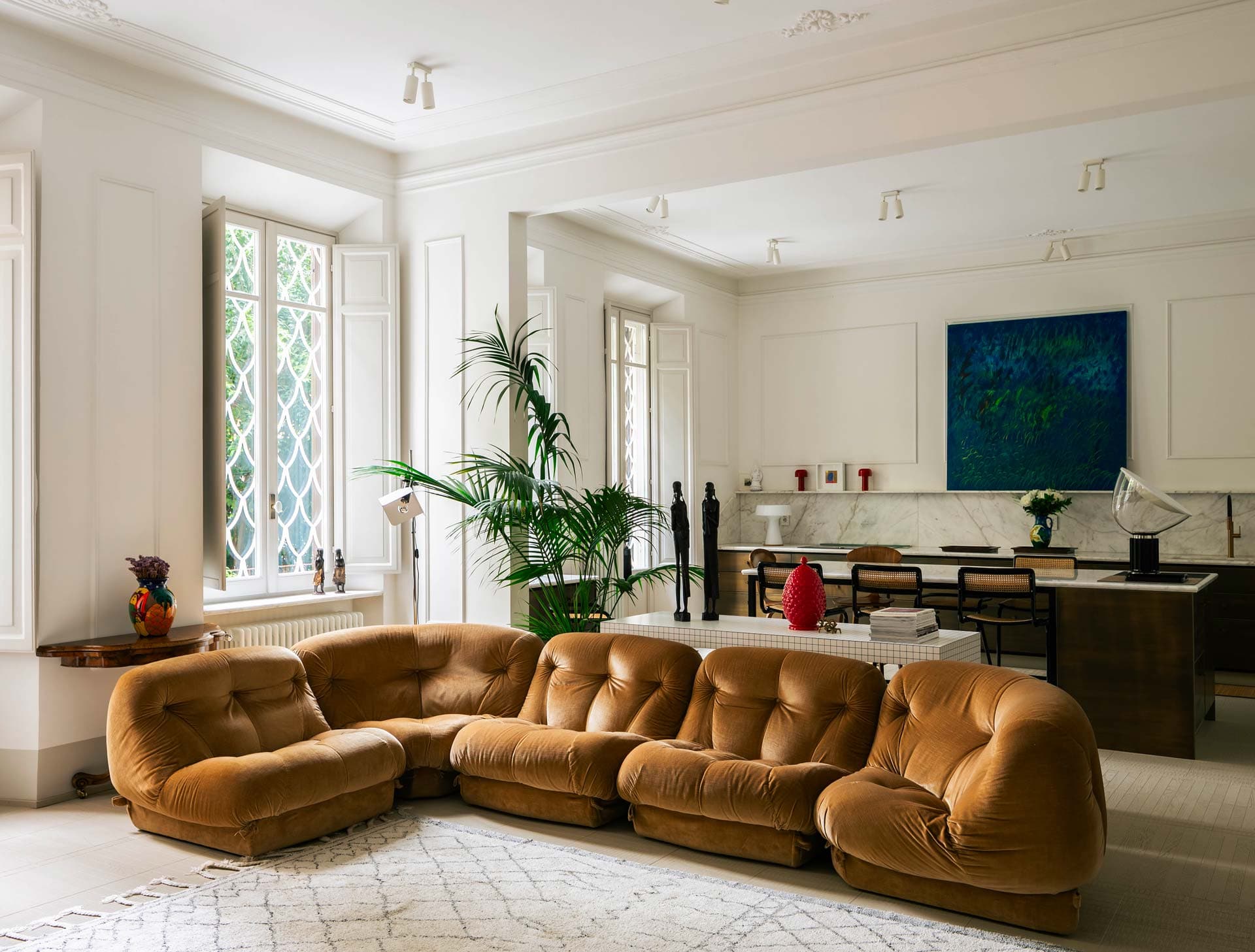 Stylish living room with a brown sectional sofa, large windows, indoor plants, a dining area, and abstract wall art.