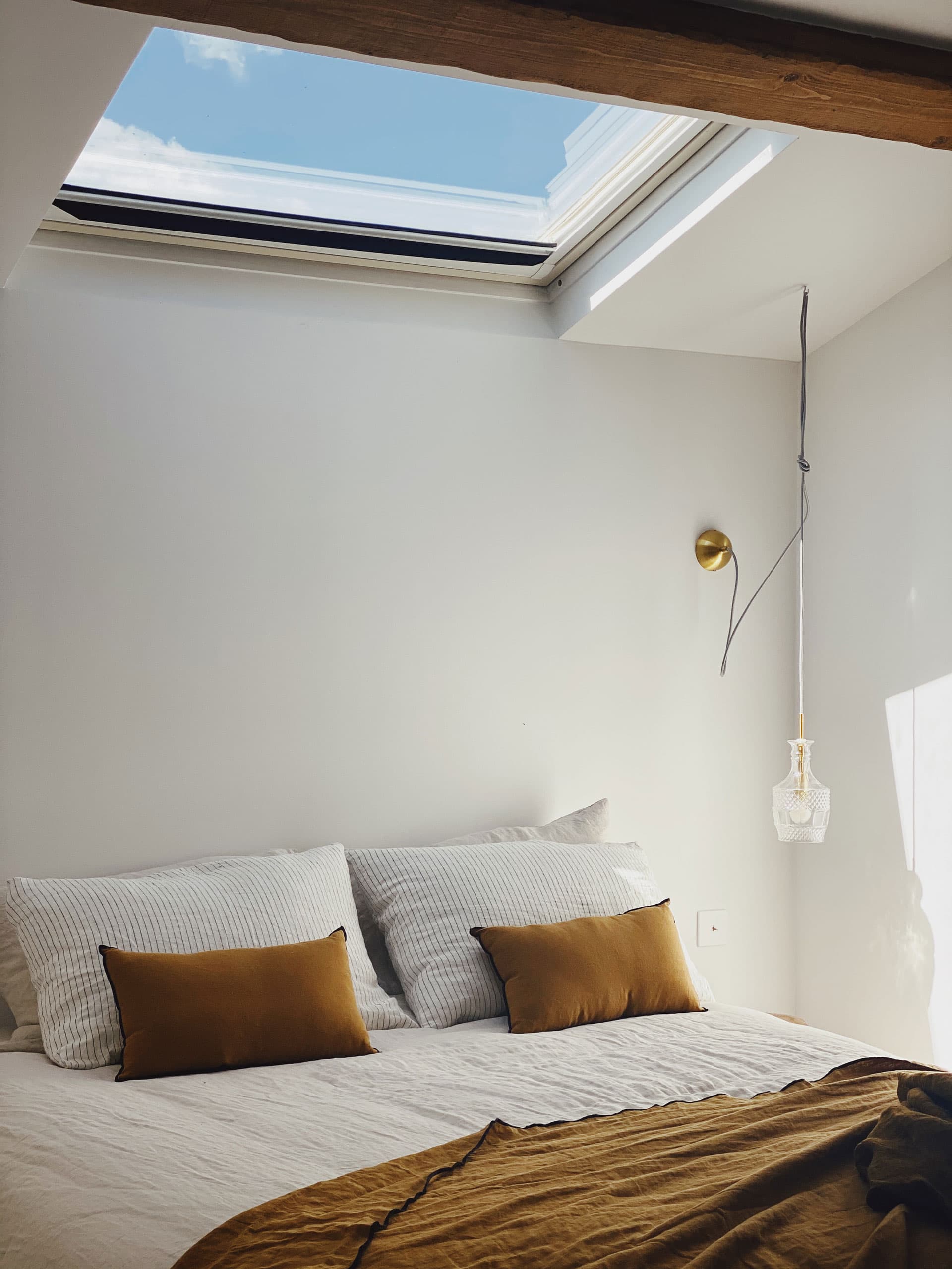 Cozy bedroom with a skylight, white bedding, mustard pillows, a wooden beam, and a hanging light fixture.