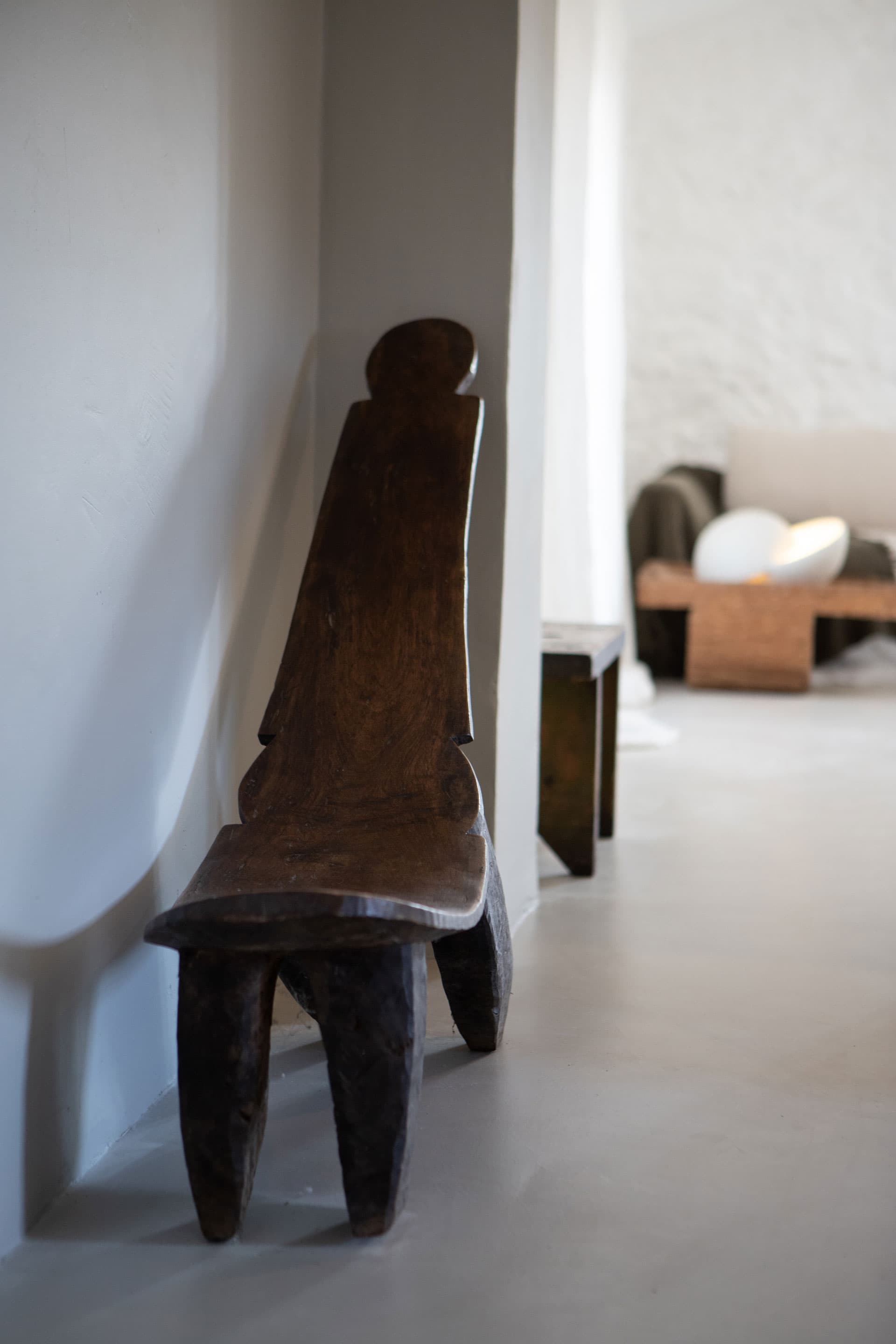 A wooden chair, carved backrest leans in a minimalist room with a soft-lit atmosphere.