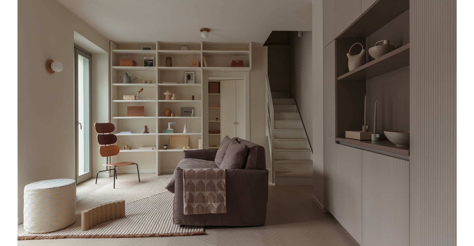 Minimalist living room with a gray sofa, modern chair, open shelving, and neutral decor. A staircase leads upstairs. Soft, natural lighting.