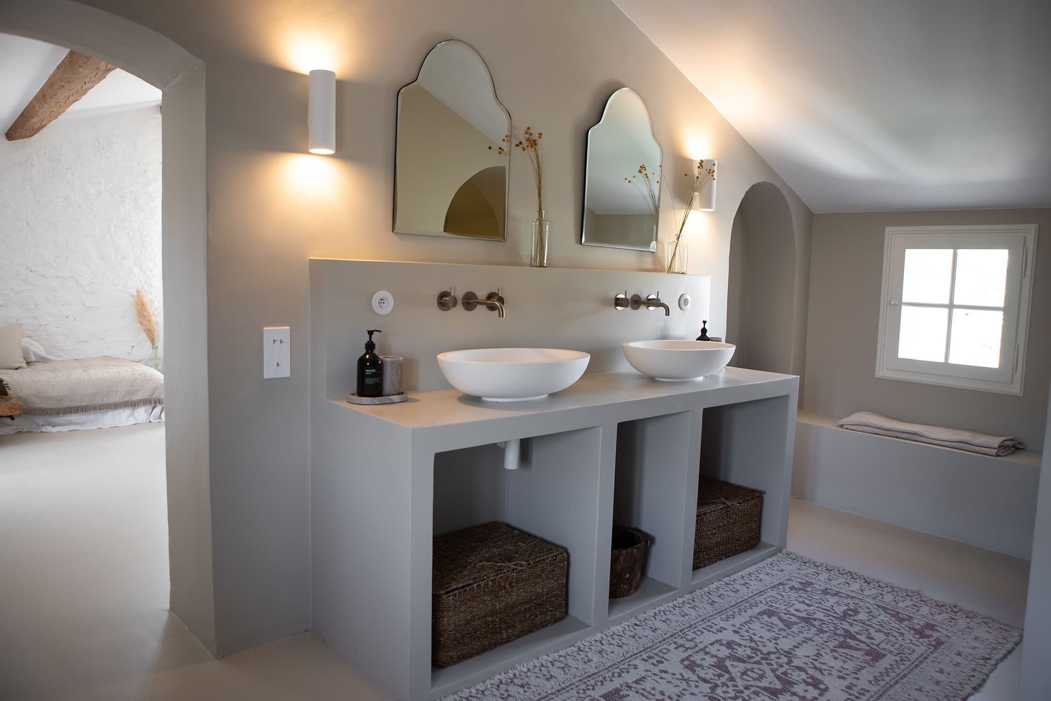 Modern bathroom with dual sinks, arched mirrors, and wall-mounted faucets. Wicker baskets beneath and a window illuminating the space.