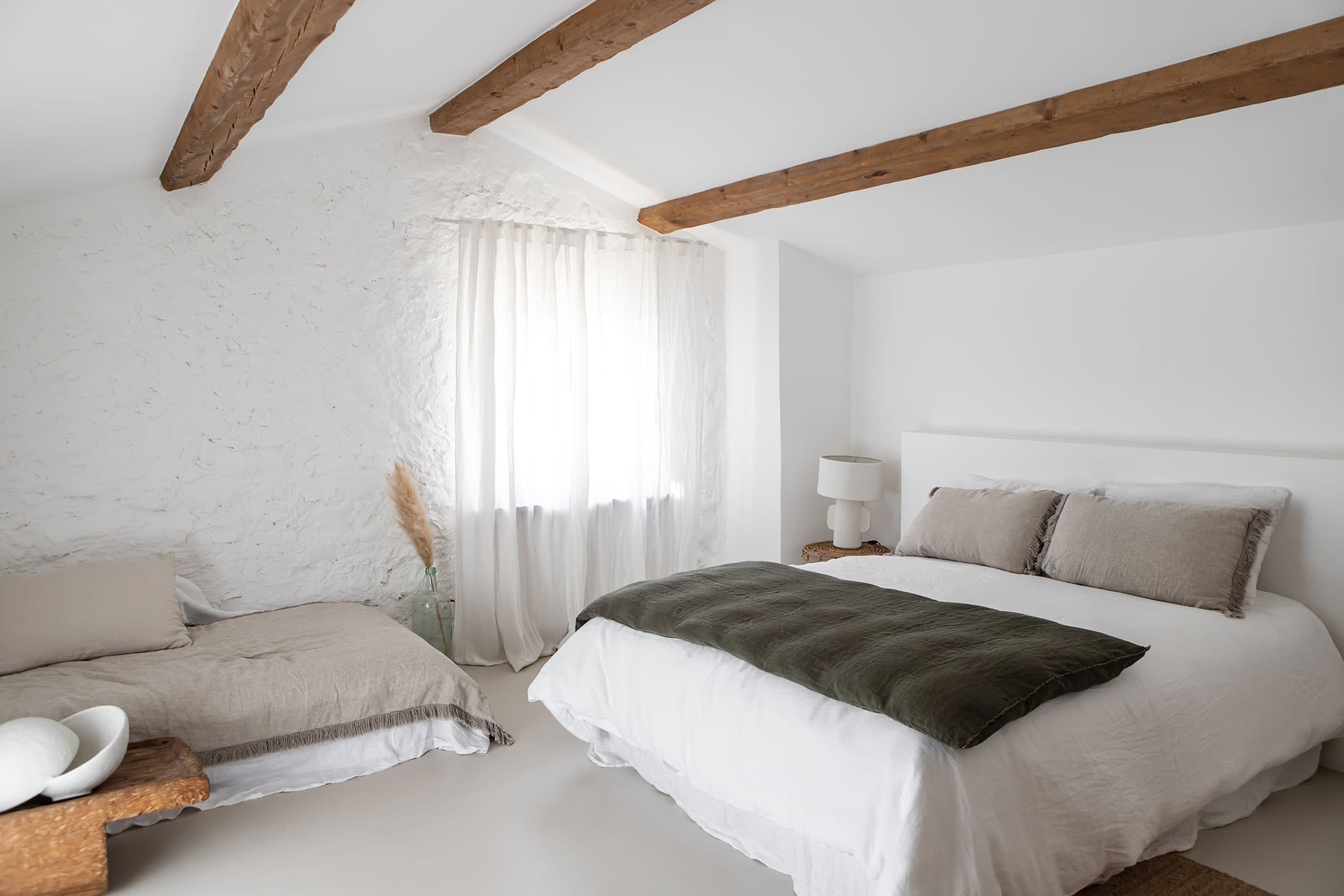 Minimalist bedroom with white walls, wooden beams, a large bed with gray and white bedding, a small side table, and soft natural light through curtains.