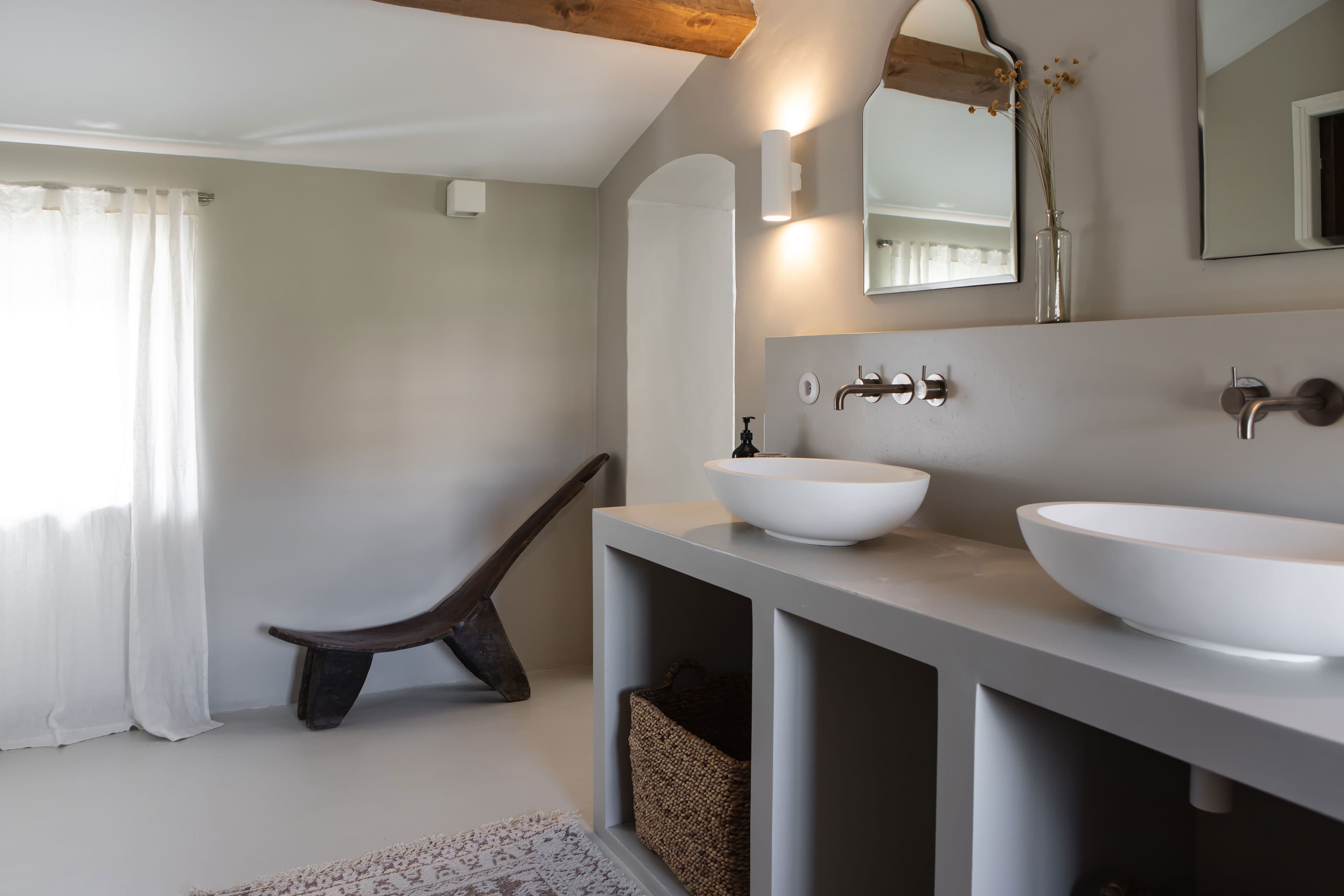 Minimalist bathroom with two vessel sinks, wall-mounted faucets, a wooden chair, and a large window with sheer curtains.