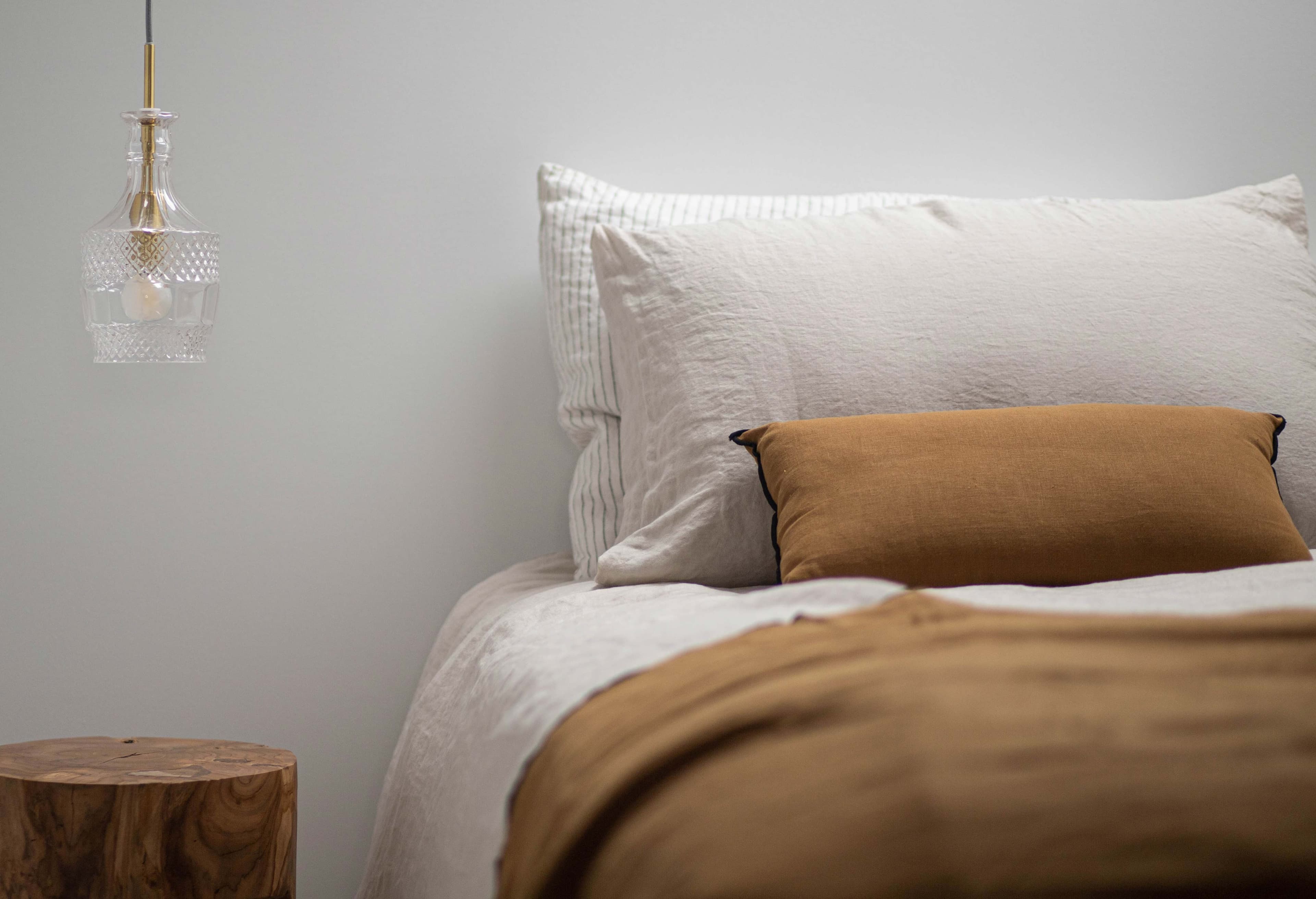 Minimalist bedroom with a neatly made bed featuring white and brown bedding, a wooden side table, and a hanging glass pendant light.
