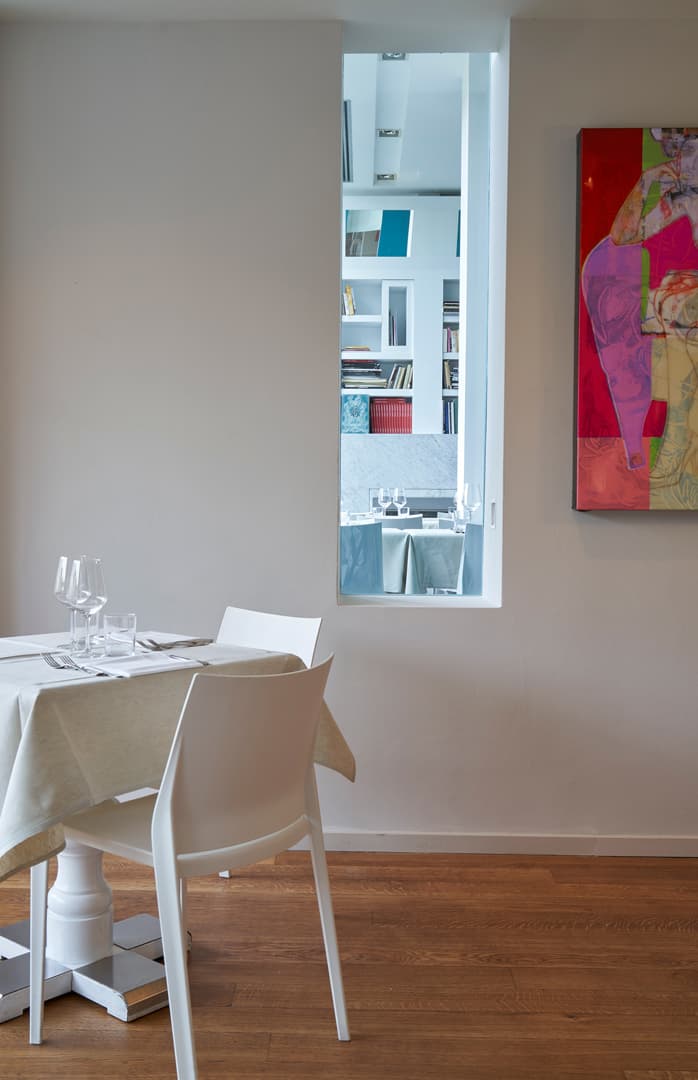 Minimalist dining area with a white table and chairs, a colorful abstract painting on the wall, and a window revealing a bookshelf.