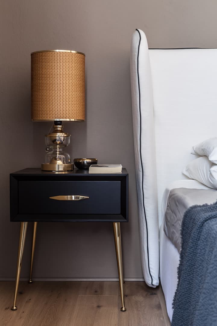 A modern bedroom with a black nightstand, gold lamp, and a white bed with a gray blanket, set against a taupe wall.