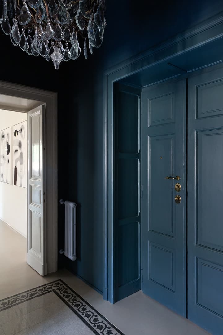 Elegant hallway with dark blue walls, ornate chandelier, and a closed blue door. White door and modern artwork visible in the background.