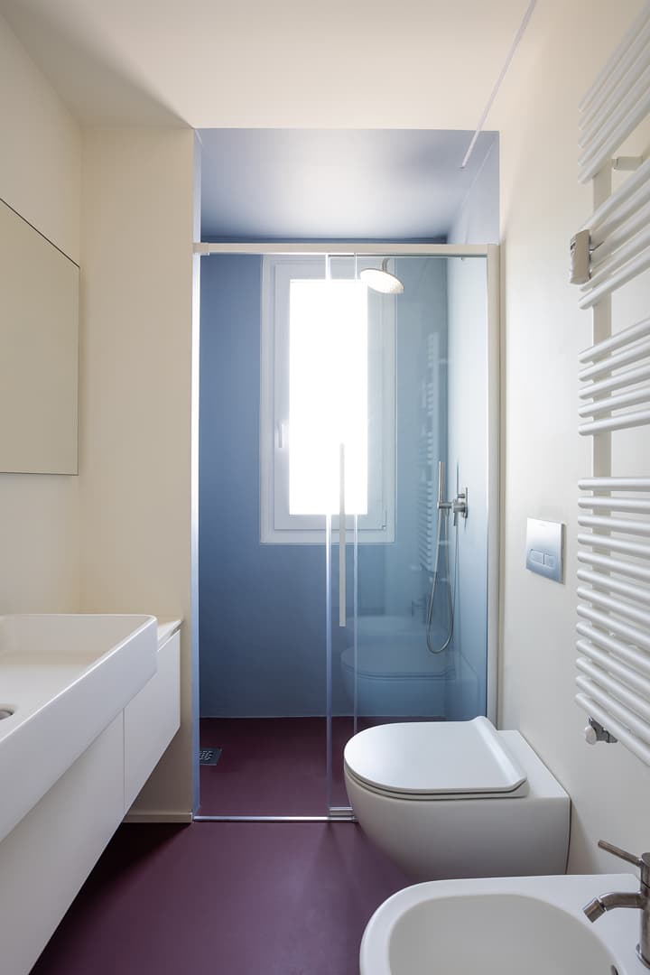 Minimalist bathroom with purple floor, glass shower and white fixtures. Natural light from a window brightens the space.