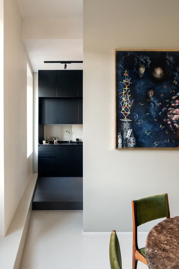 Modern interior with a black kitchen, abstract painting on a beige wall, and a green chair next to a round table with a marble surface.
