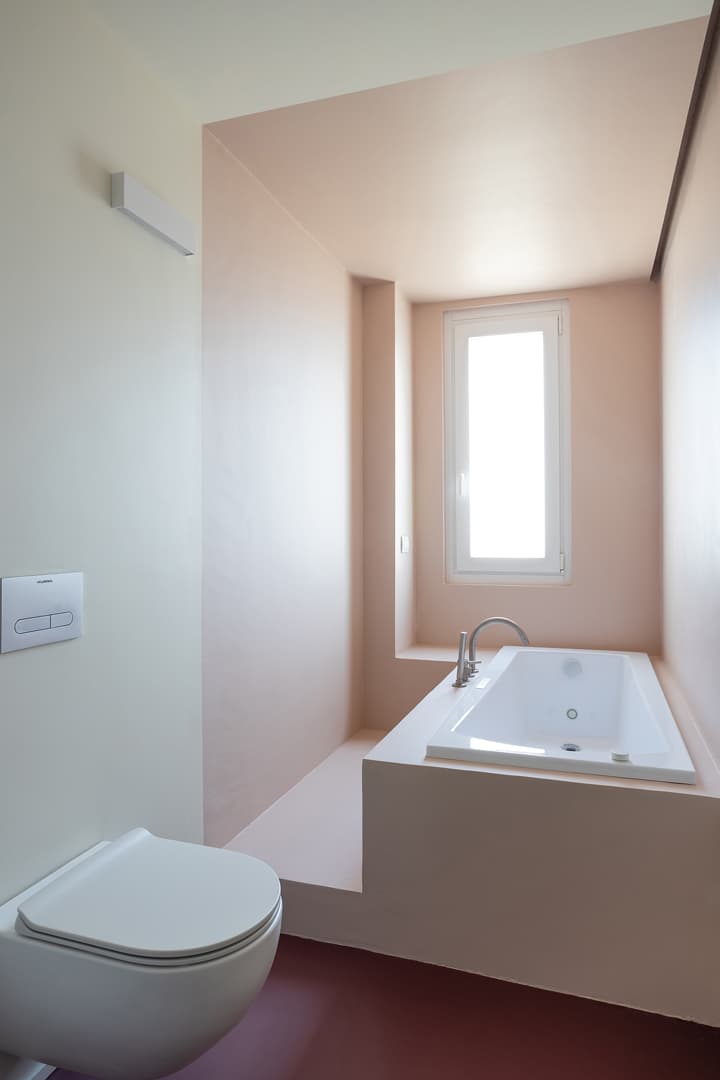 Minimalist bathroom with a white wall-mounted toilet and a built-in bathtub under a tall window, featuring soft pink and white tones.